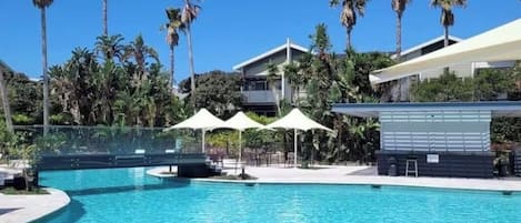 En swimmingpool med palmer, et overdækket opholdsområde og en bygning med moderne arkitektur.