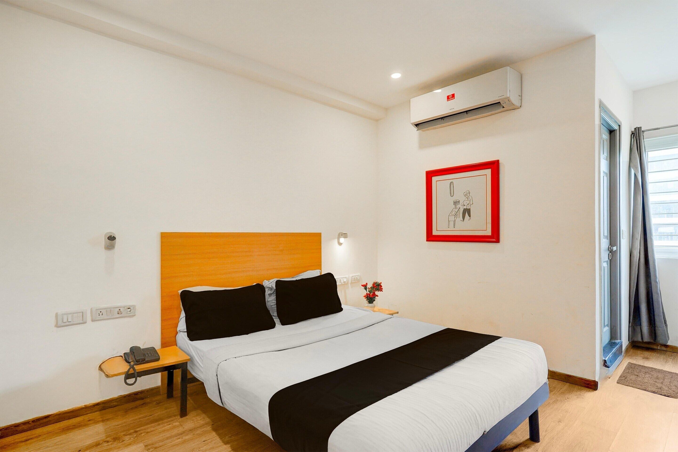 A hotel room with a bed, bedside table, phone, wall-mounted air conditioner, and a picture frame.