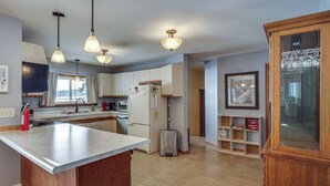 Una cucina con isola centrale, frigorifero, lavello e un cestino per i rifiuti. Sono presenti mobili a legno e una finestra con tende.