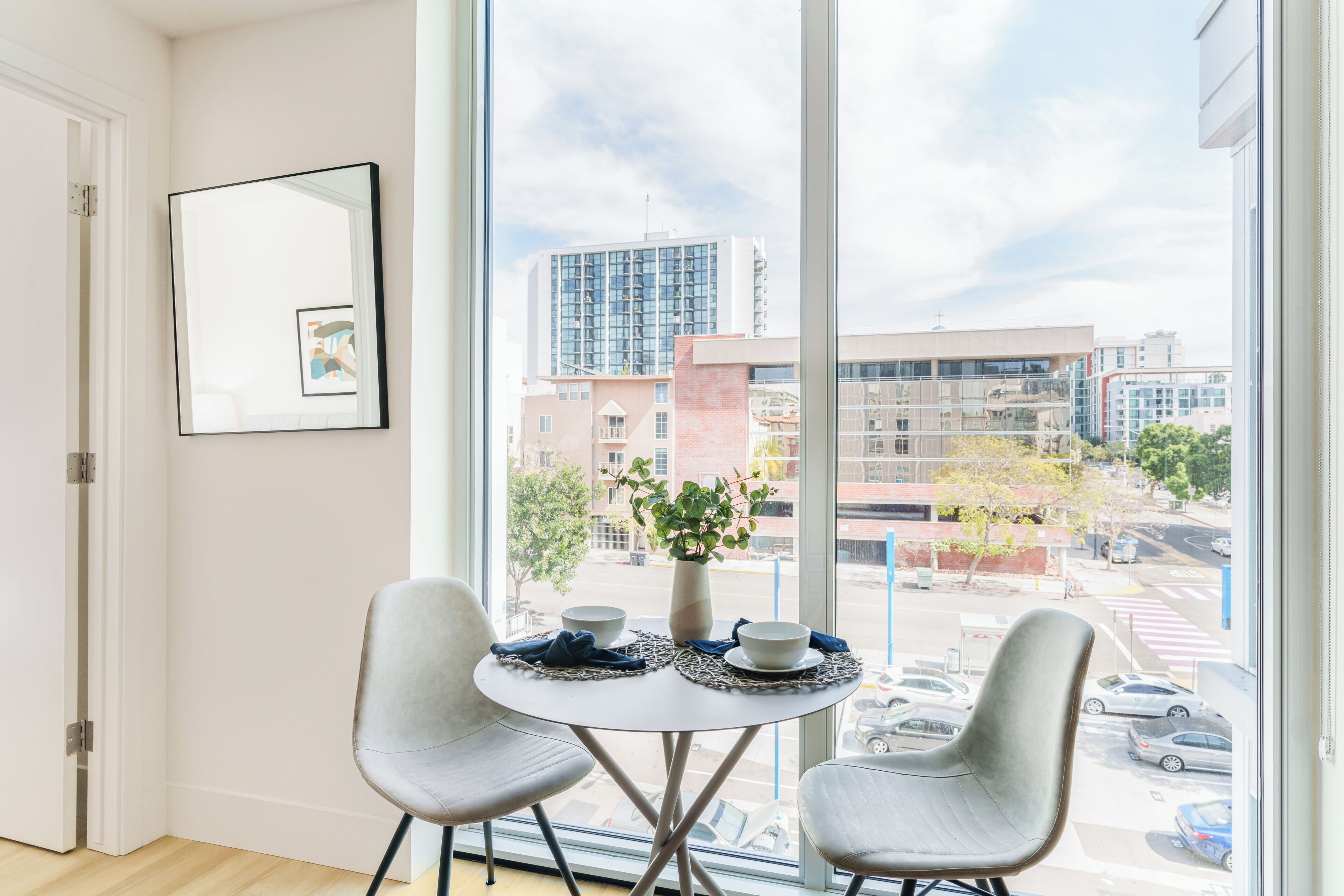 A modern room with a large window, a dining table with two chairs, a vase with flowers, and a framed picture on the wall.