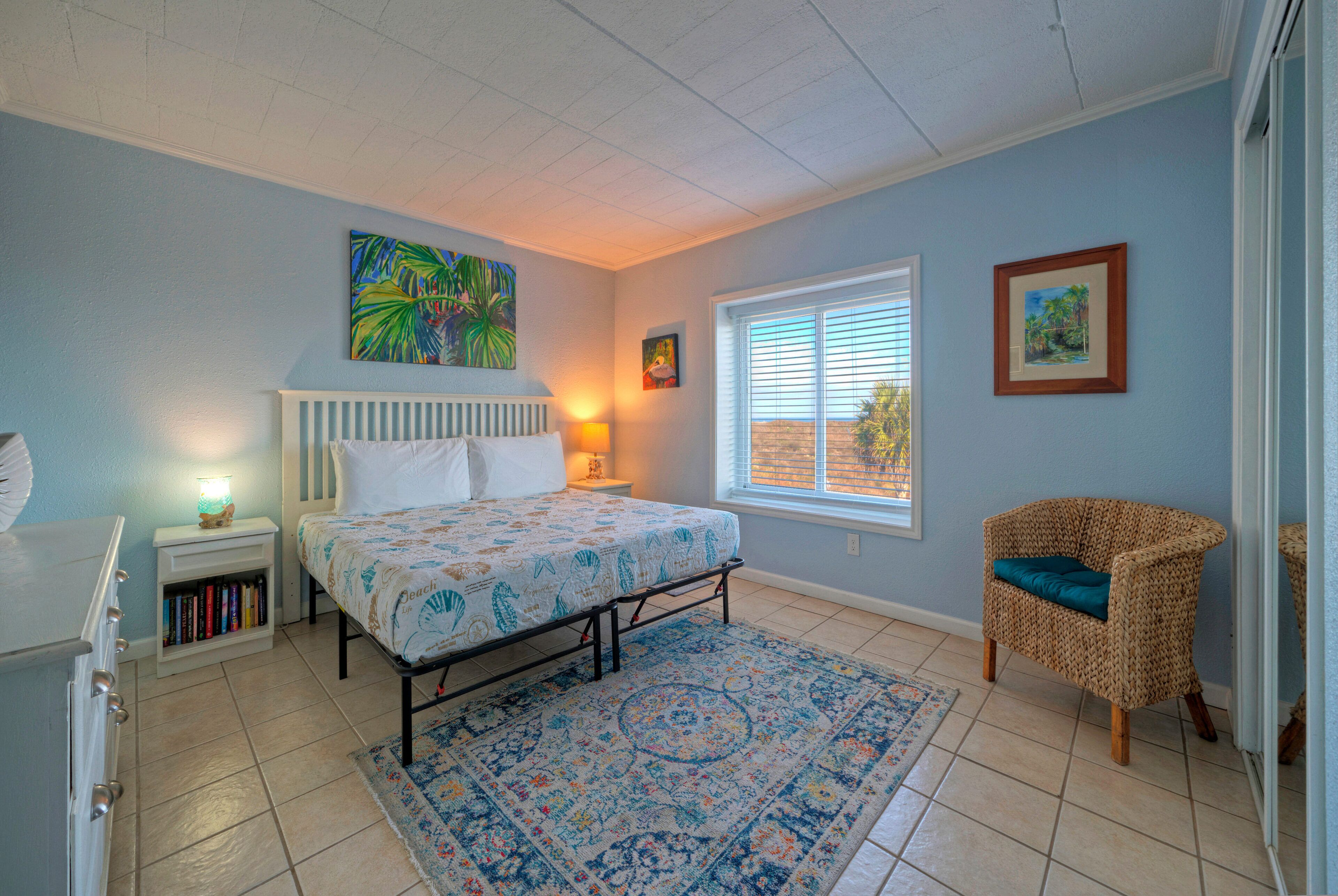 A bedroom with a bed, a nightstand, a chair, a window, and artwork on the walls.