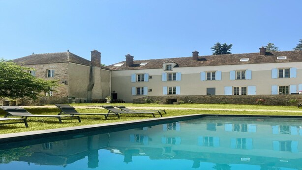 Ein Schwimmbecken mit Liegestühlen, ein Steinhaus mit vielen Fenstern und ein klarer, blauer Himmel.