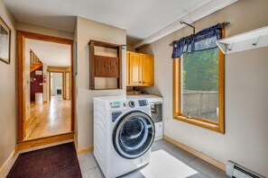 A laundry room with a front-loading washer and dryer, wooden cabinets, a window with a view, and a door leading to another room. - Explore the White Mountains! Spacious Rural Home (Thornton)