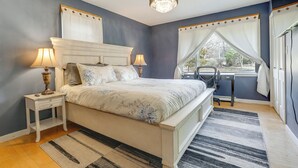 Quarto com cama, duas mesas de cabeceira com luminárias, escrivaninha, cadeira, janela com cortinas e lustre.