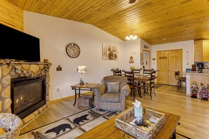 A living room with a stone fireplace, a television, a clock, and a dining area with wooden furniture. - Rocky Mtn-View Retreat! Walk to Dtwn Grand Lake (Grand Lake)