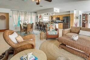 A spacious living room with wicker furniture, a dining area with chairs, a kitchen with wooden cabinets, and a ceiling fan.