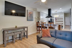 A modern living room with a grey sofa, wooden dining table, and chairs. A kitchen with white cabinets and a black range hood is visible in the background.