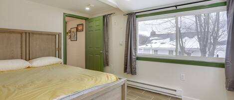 A bedroom with a bed, a green door, a window with curtains, and a view of a house.