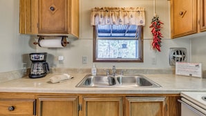 A kitchen with wooden cabinets, a stainless steel sink, a coffee maker, and a window with a view of trees. - Near Grindstone Lake & Family Fun! Cozy Cabin (Ruidoso)
