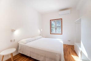 Ein Schlafzimmer mit einem Bett, einem kleinen runden Tisch, einem Fenster mit Holzrahmen und einer Klimaanlage an der Wand.