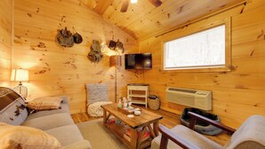 En mysig stuga med träpanel i taket, en tv som hänger på väggen, ett fönster med persienner, ett träbord och en soffa.