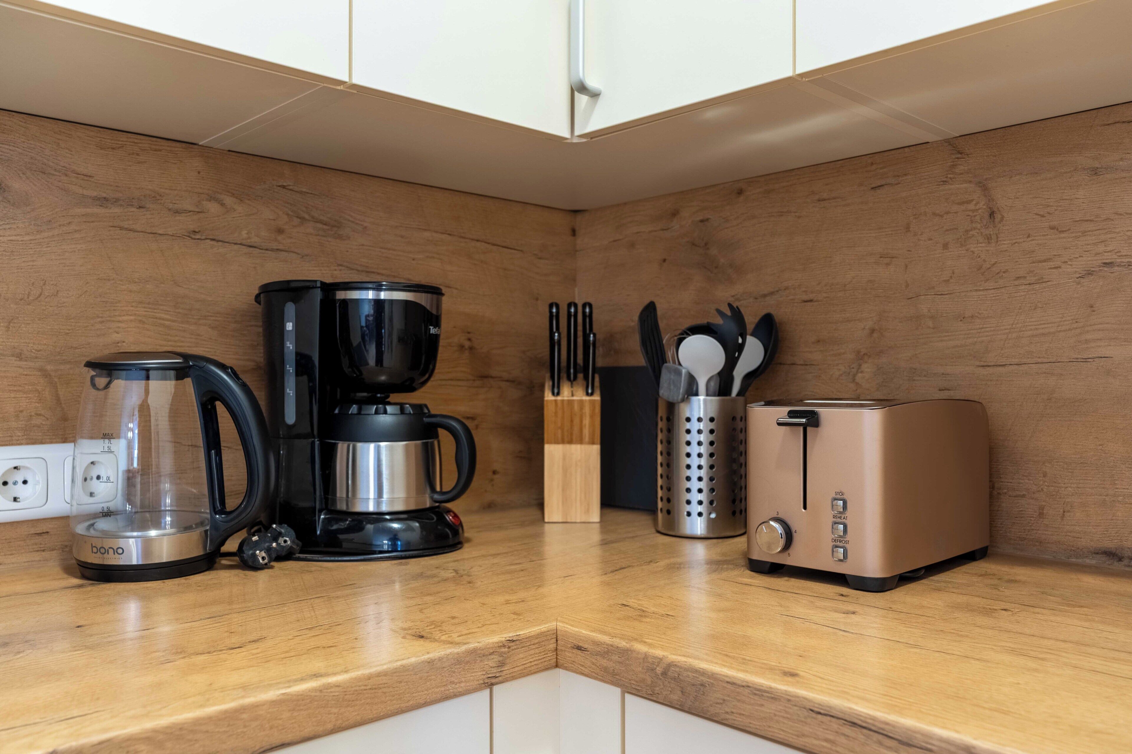 Una cocina con cafetera, tostadora, bloque de cuchillos y una jarra de vidrio sobre una encimera de madera.
