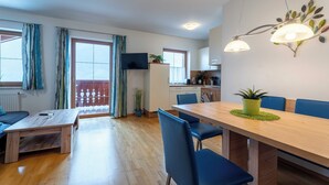 Ein modernes Wohnzimmer mit einem Holzesstisch, blauen Stühlen, einem Fernseher und einer Küchenzeile mit Schränken und einer Pflanze.