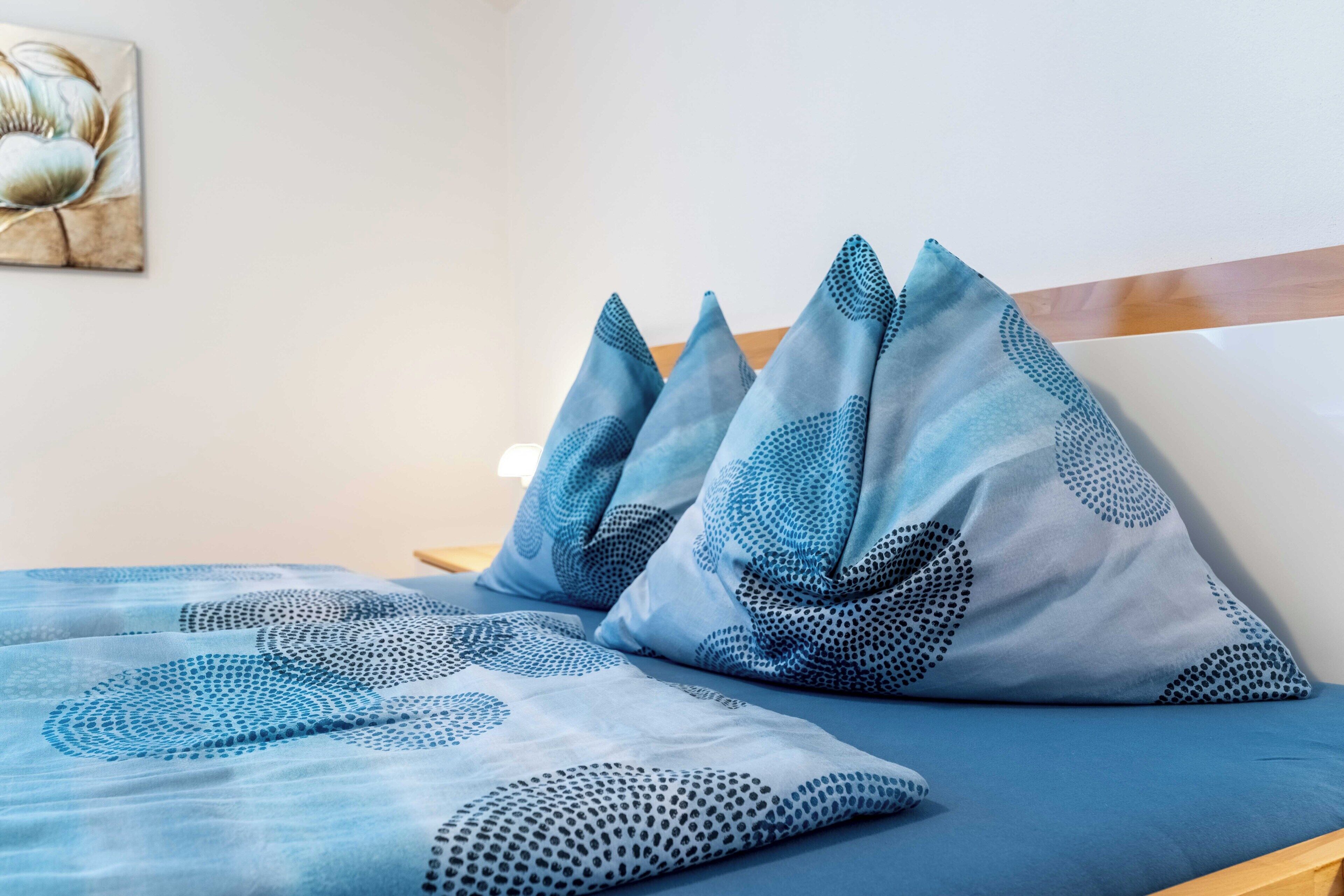 A bed with blue bedding and pillows, a wooden headboard, and a wall with a framed artwork.