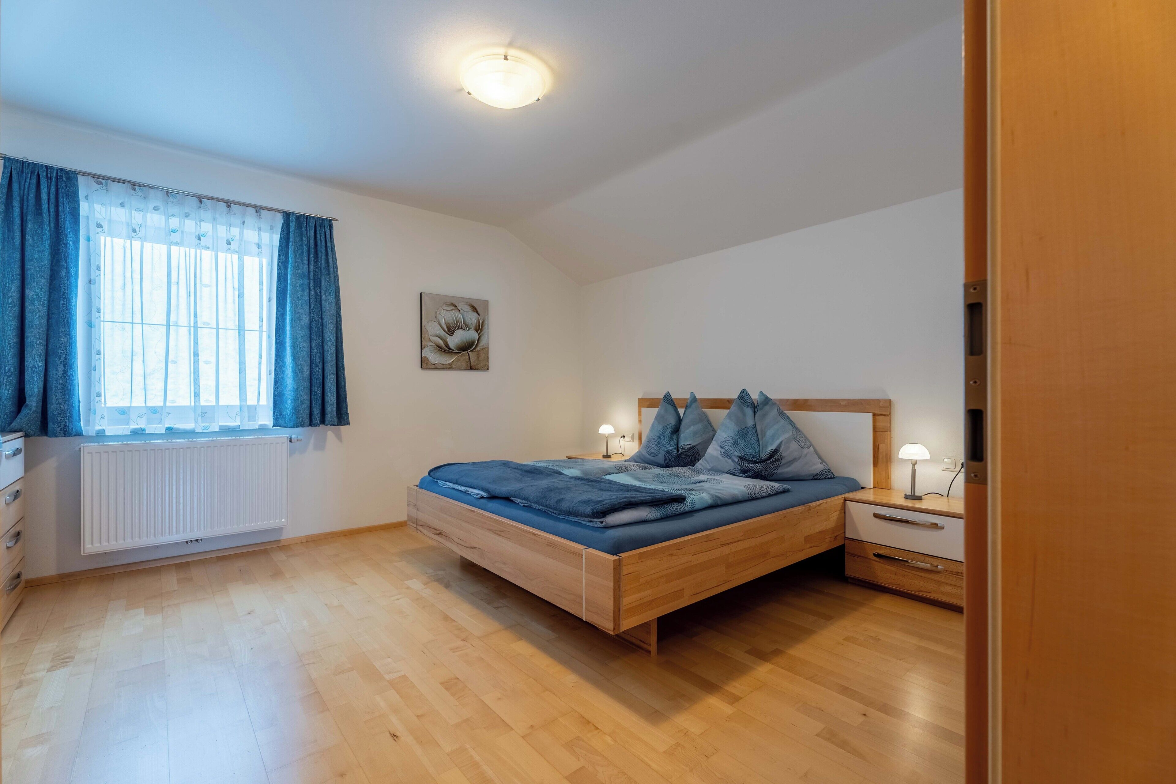 Ein Schlafzimmer mit einem hölzernen Bett, einem Nachttisch mit Lampe, einem Fenster mit Vorhängen und einer Heizung.