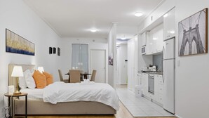 Quarto de hotel moderno com cama, mesa de cabeceira, abajur e vista para a cozinha adjacente.