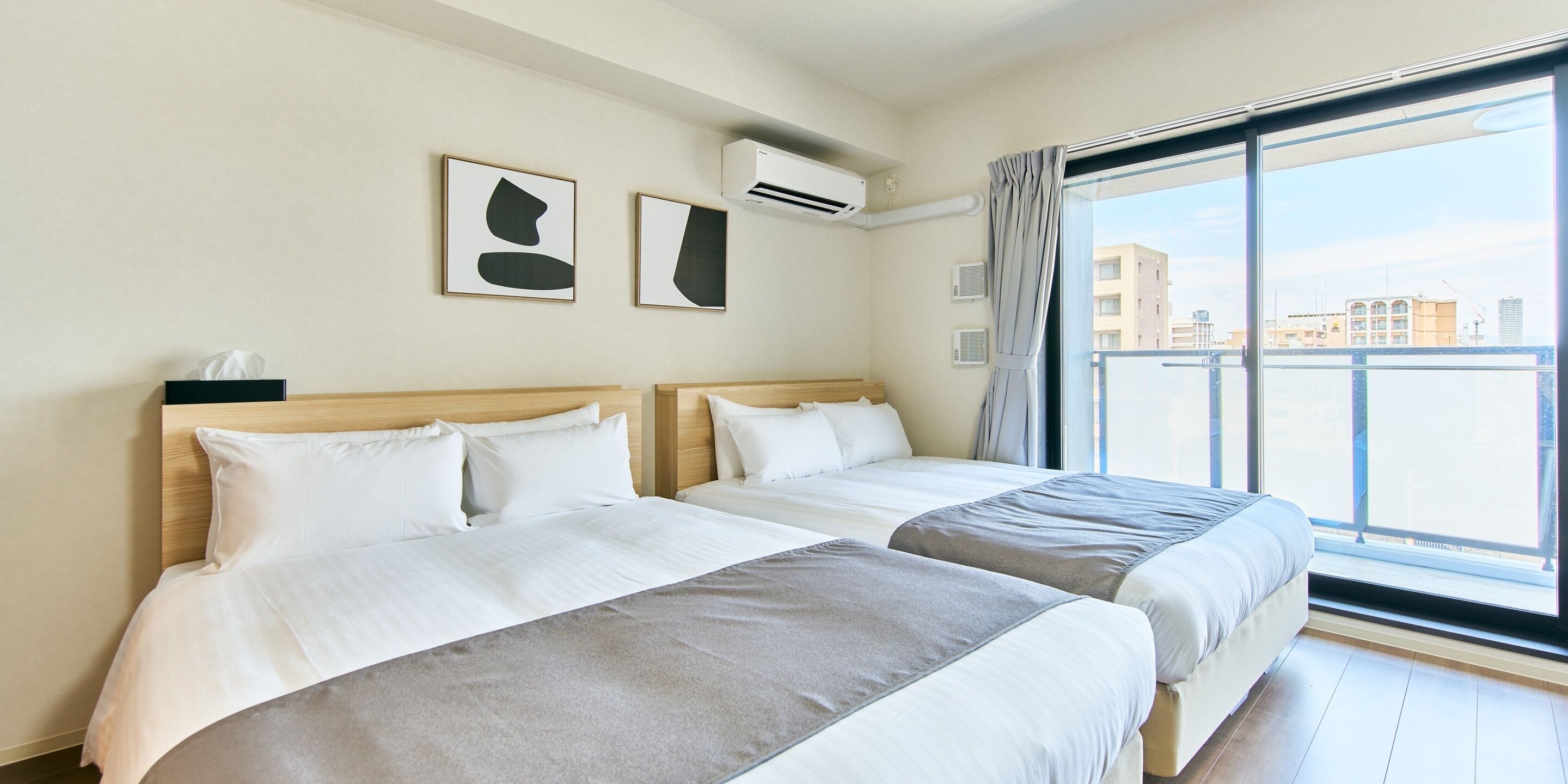 A hotel room with two beds, a large window with a view of buildings, and an air conditioning unit on the wall.