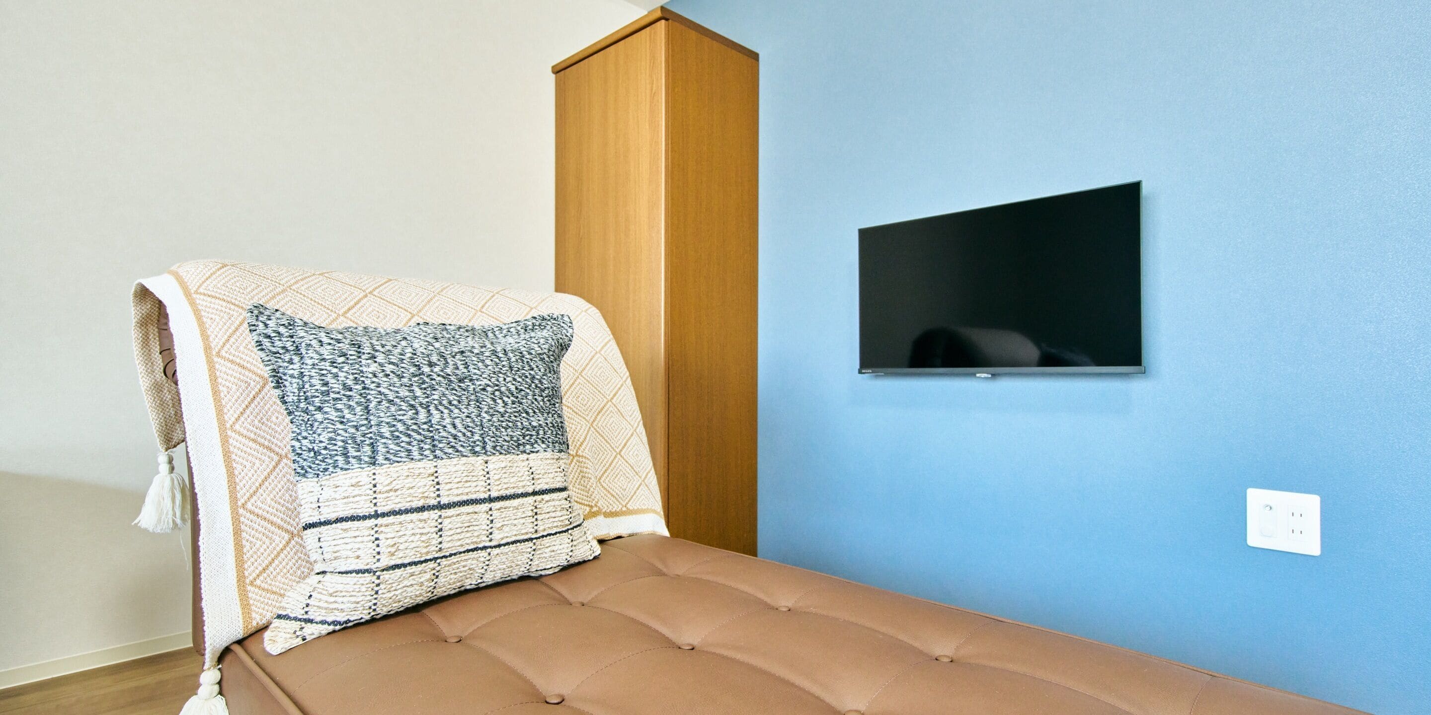 A single bed with a brown headboard and a patterned blanket, a wooden wardrobe, and a small TV mounted on a blue wall.