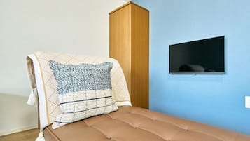 A single bed with a brown headboard and a patterned blanket, a wooden wardrobe, and a small TV mounted on a blue wall.
