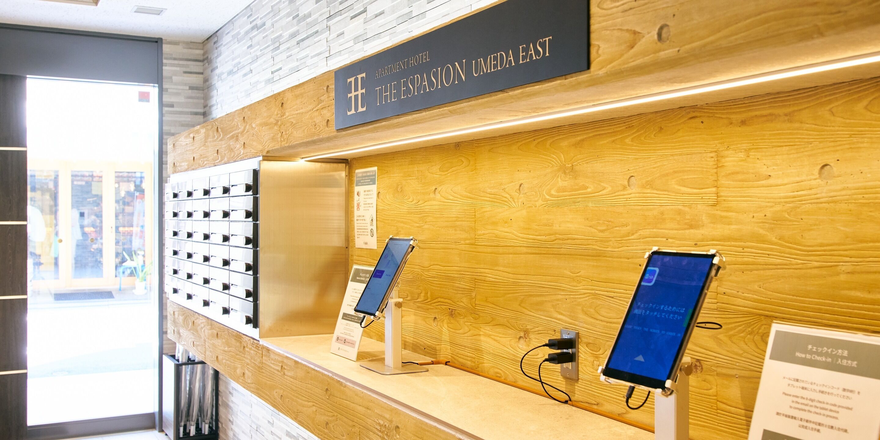 A reception desk with a sign for 'The Espasion Umeda East' hotel, a digital check-in kiosk, and a wall-mounted information board.