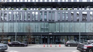 Un bâtiment moderne doté de grandes fenêtres, d’une entrée noire et de voitures garées devant.