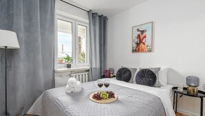 Une chambre à coucher avec un lit, une table de chevet avec une lampe, une fenêtre avec des rideaux, un tableau encadré au mur et une assiette de fruits sur le lit.