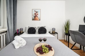 A neatly made bed with towels, a fruit platter, and wine glasses, a small table with books and a lamp, and a potted plant. - Cozy Apartment with balcony near Museum (Warsaw)