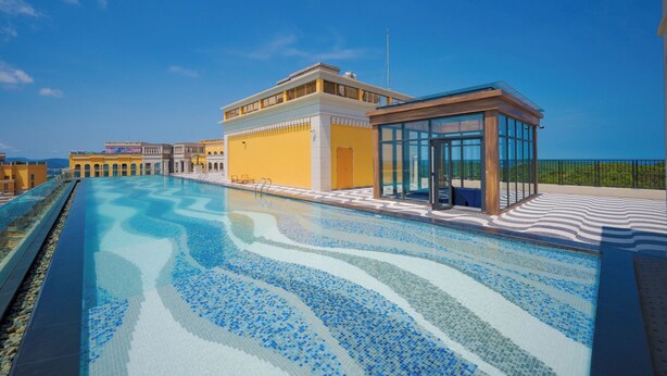 Uma piscina com padrão ondulado, um edifício amarelo e uma estrutura com paredes de vidro.