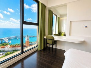 Habitación de hotel con un ventanal grande, cama, escritorio con silla y vistas a un paisaje urbano costero.