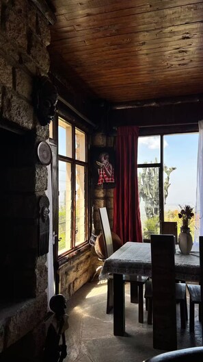Un interior rústico con paredes de piedra, vigas de madera, una mesa de comedor con sillas y vistas a cactus a través de una ventana.