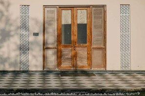 Puerta de madera con paneles de vidrio, flanqueada por celosías metálicas decorativas, sobre un muro liso con suelo de tablones.