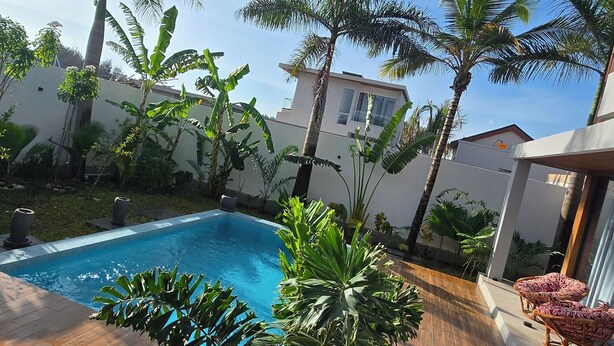 A swimming pool surrounded by wooden decking, palm trees, and a white building with windows. - Villa Casa Casar in Paje Zanzibar for rent (Sansibar)