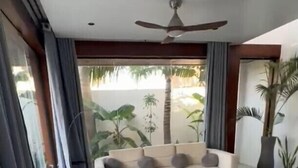 A modern living room with a sofa, ottoman, and coffee table, large windows with curtains, and a ceiling fan. - Villa Casa Casar in Paje Zanzibar for rent (Sansibar)