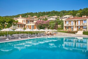 A swimming pool with lounge chairs and umbrellas, surrounded by buildings with balconies and a hillside in the background. - Apartment on Lake Garda (Torri del Benaco VR)