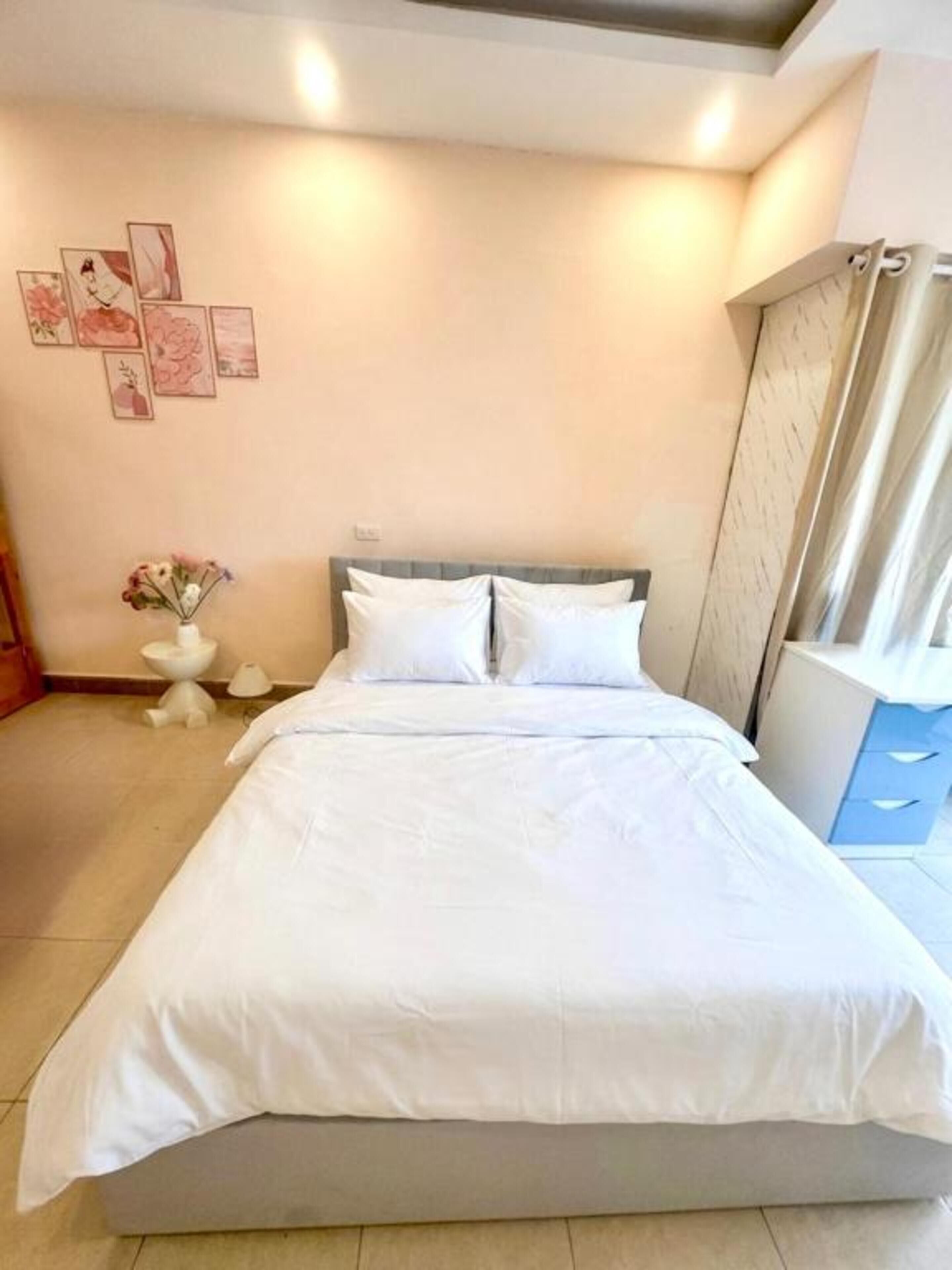 A neatly made bed in a hotel room with a headboard, a small bedside table with a vase of flowers, and a wall with framed pictures.