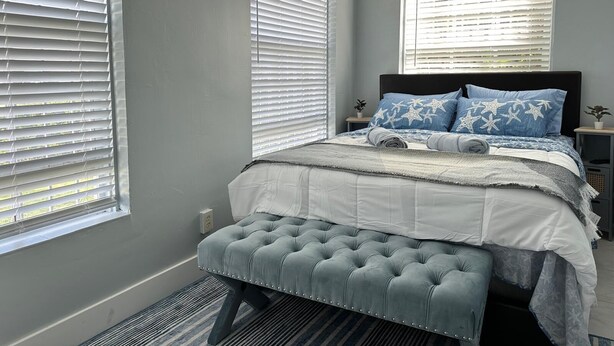 Quarto com cama, um banquinho e duas janelas com persianas.