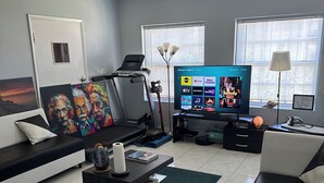 Uma sala de estar moderna com uma esteira, uma televisão de tela plana e uma grande janela com persianas.