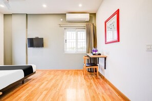Habitación de hotel con cama, escritorio y silla, televisor, equipo de aire acondicionado, ventana con cortinas e imagen enmarcada en la pared.
