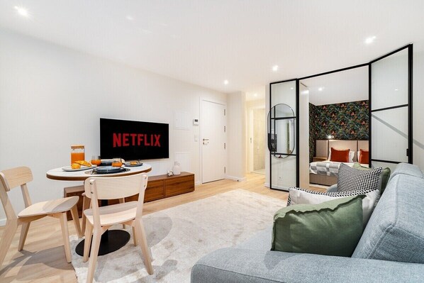 Ein modernes Wohnzimmer mit Flachbildfernseher, einem Essbereich mit Tisch und Stühlen sowie einer gemütlichen Sitzecke mit Sofa und Kissen.
