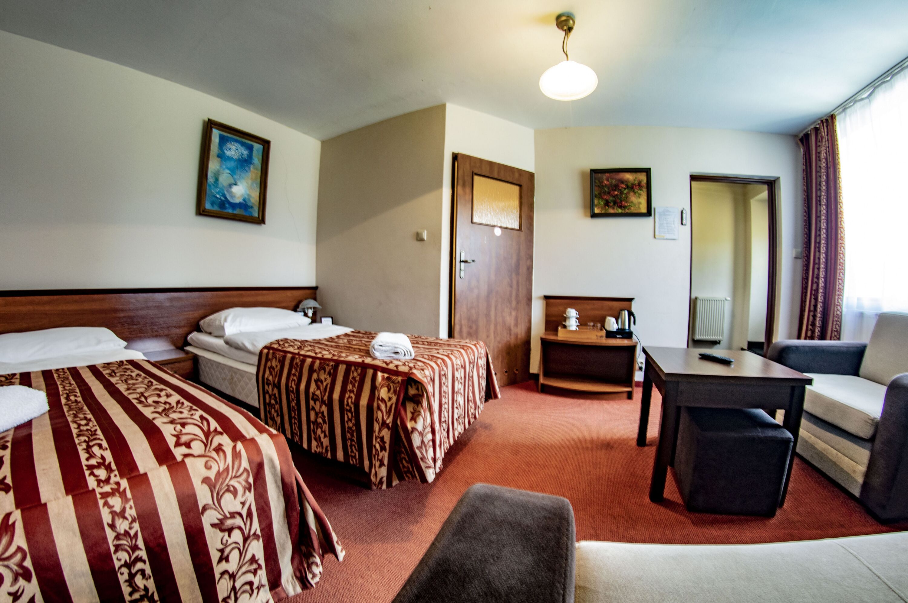 A hotel room with two beds, a desk, a chair, and a small table with a kettle and cups.