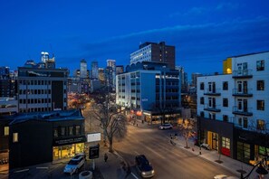 A cityscape at dusk with modern buildings, a clinic, and a street with cars and pedestrians. - Mins to Saddledome | UG Parking | 2 Bed | Central (Calgary)