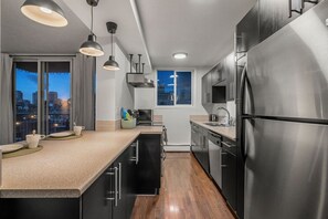 A modern kitchen with stainless steel appliances, a wooden floor, and a city view through large windows. - Mins to Saddledome | UG Parking | 2 Bed | Central (Calgary)