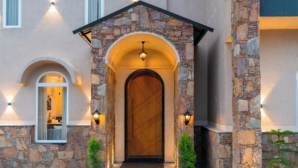 Une façade en pierre avec une porte en bois, deux plantes en pot et un luminaire au-dessus de la porte.