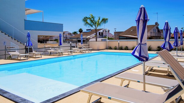 Une piscine avec des chaises longues et des parasols, un escalier et des bâtiments en arrière-plan.