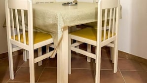 Een eettafel met tafelkleed, twee stoelen met gele kussens, en een op de muur bevestigde lichtschakelaar.
