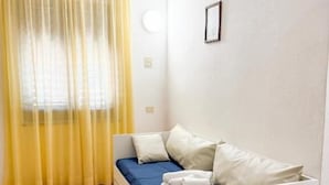 Een kleine, eenvoudige kamer met een bed, een raam met gele gordijnen, en een wandlamp.