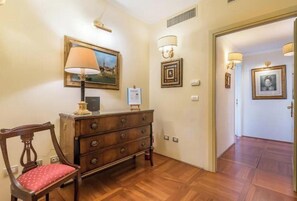 A hallway with a wooden floor, a wooden chair with a red cushion, a wooden dresser, a framed picture on the wall, and a door leading to another room. - Renaissance Elegance in the Heart of Florence (Firenze)