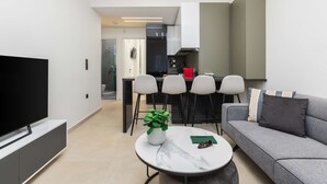 Una sala moderna con televisor de pantalla plana, una mesa de centro redonda de mármol, un sofá gris y un comedor con sillas, con una cocina al fondo.