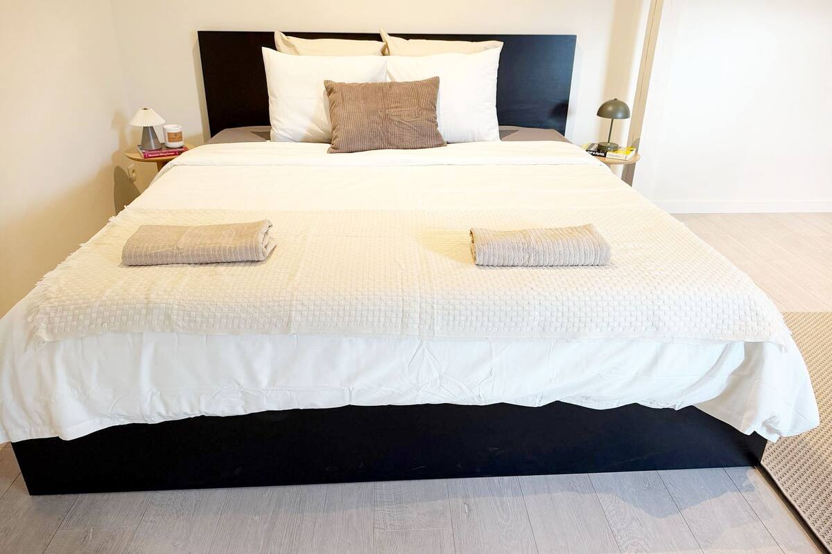 Una cama bien hecha con un cabecero negro, ropa de cama blanca y dos toallas dobladas.