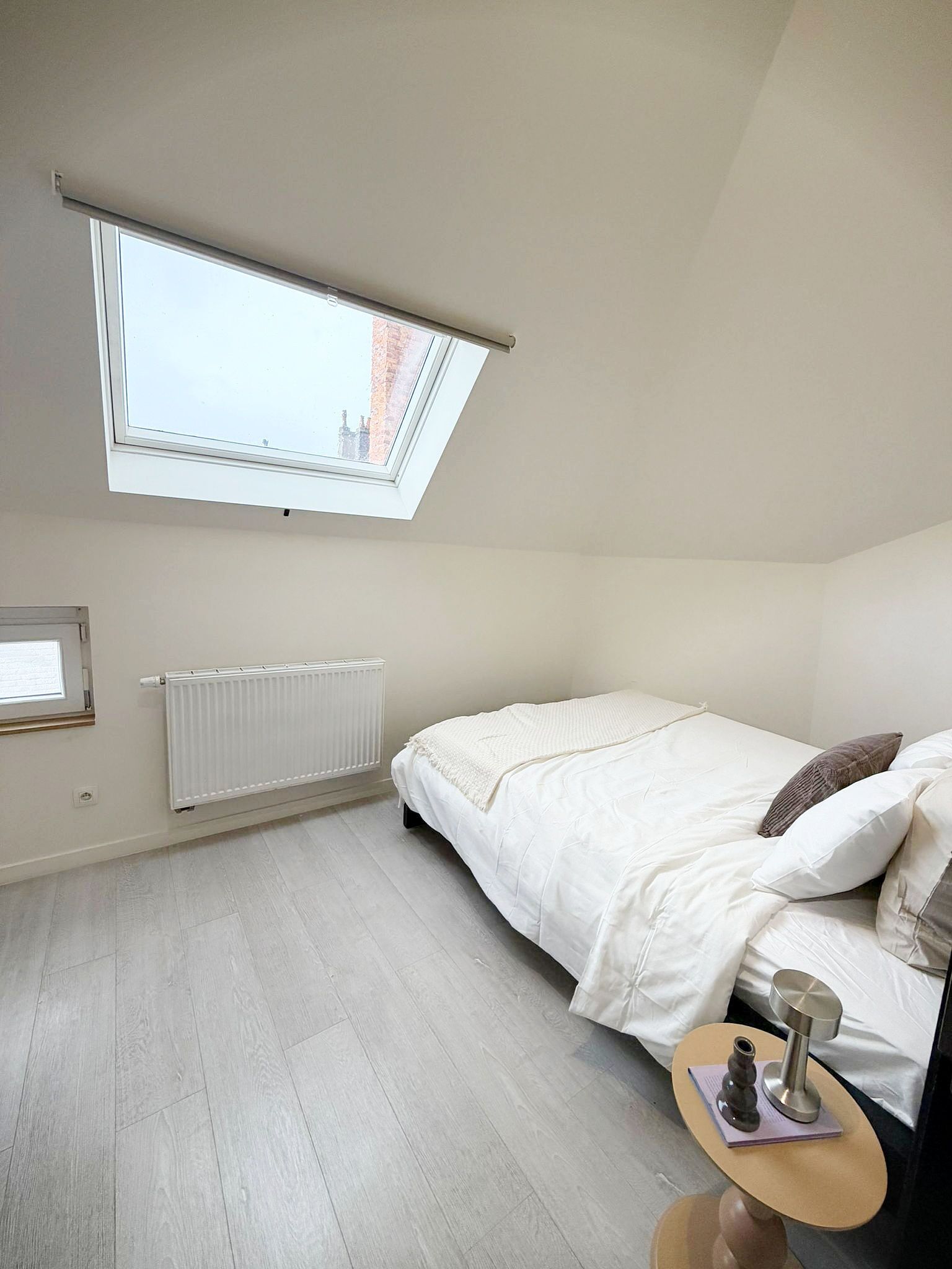 A bedroom with a skylight, a radiator, a bed with pillows, a bedside table with a vase, and a view of buildings outside.